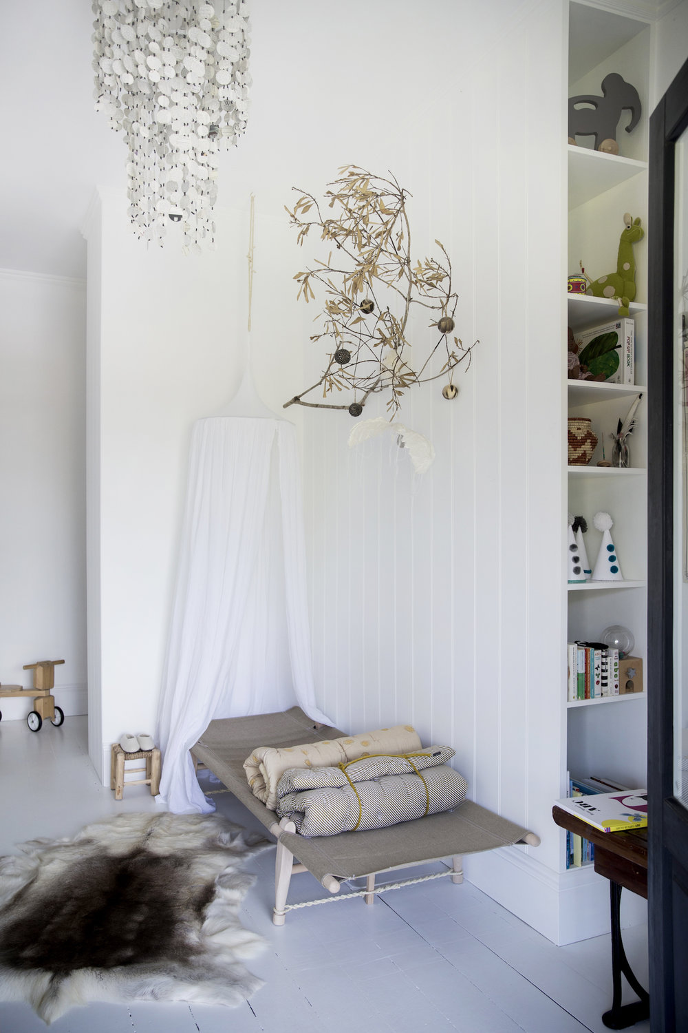Neutral-toned child's room with a daybed, decorative chandelier, white walls, and a bookshelf full of children's toys and books.