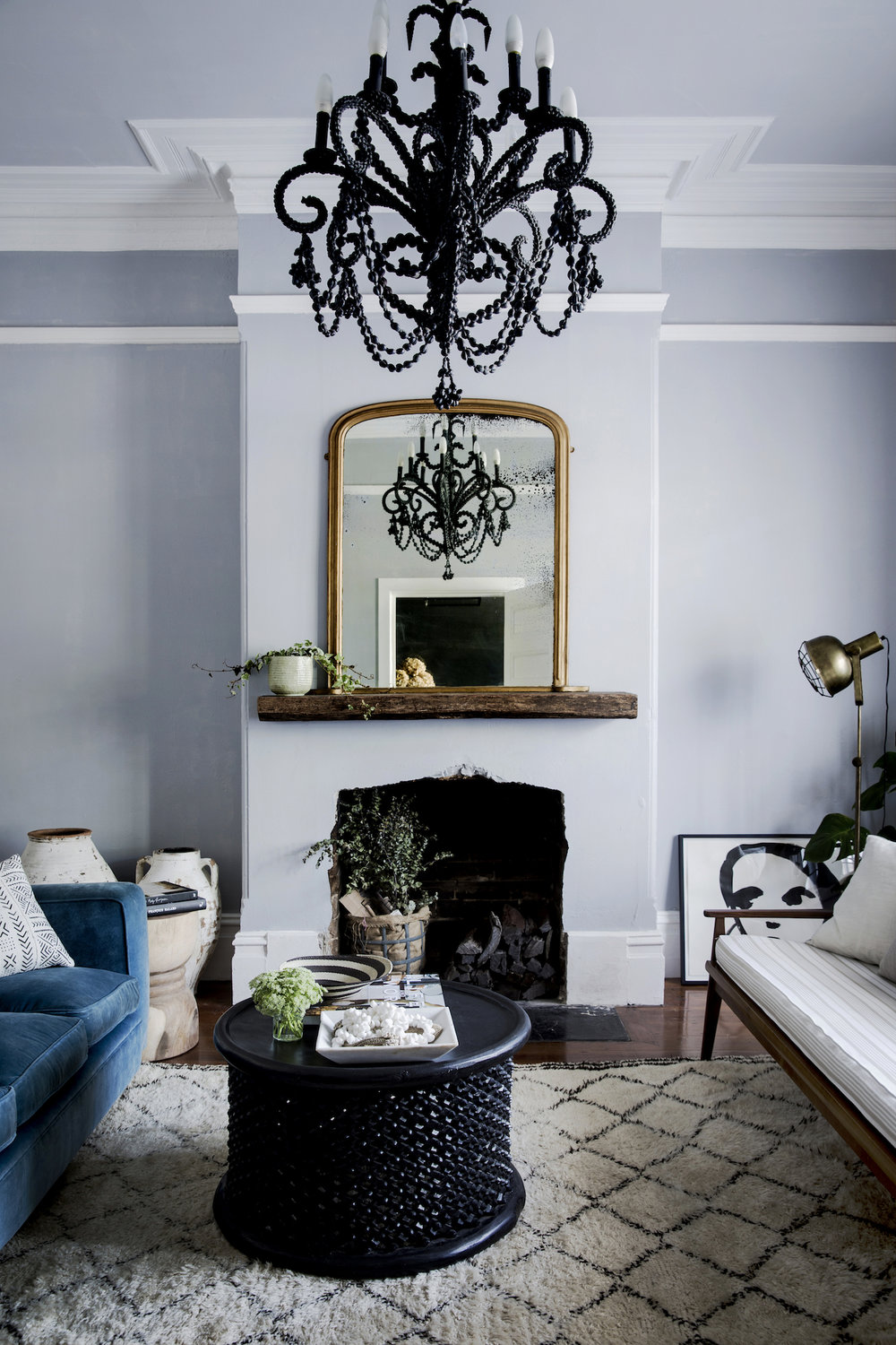 Living room interior showcasing a black chandelier, a gilded mirror over a fireplace, and a blue velvet sofa, complemented by a patterned rug.