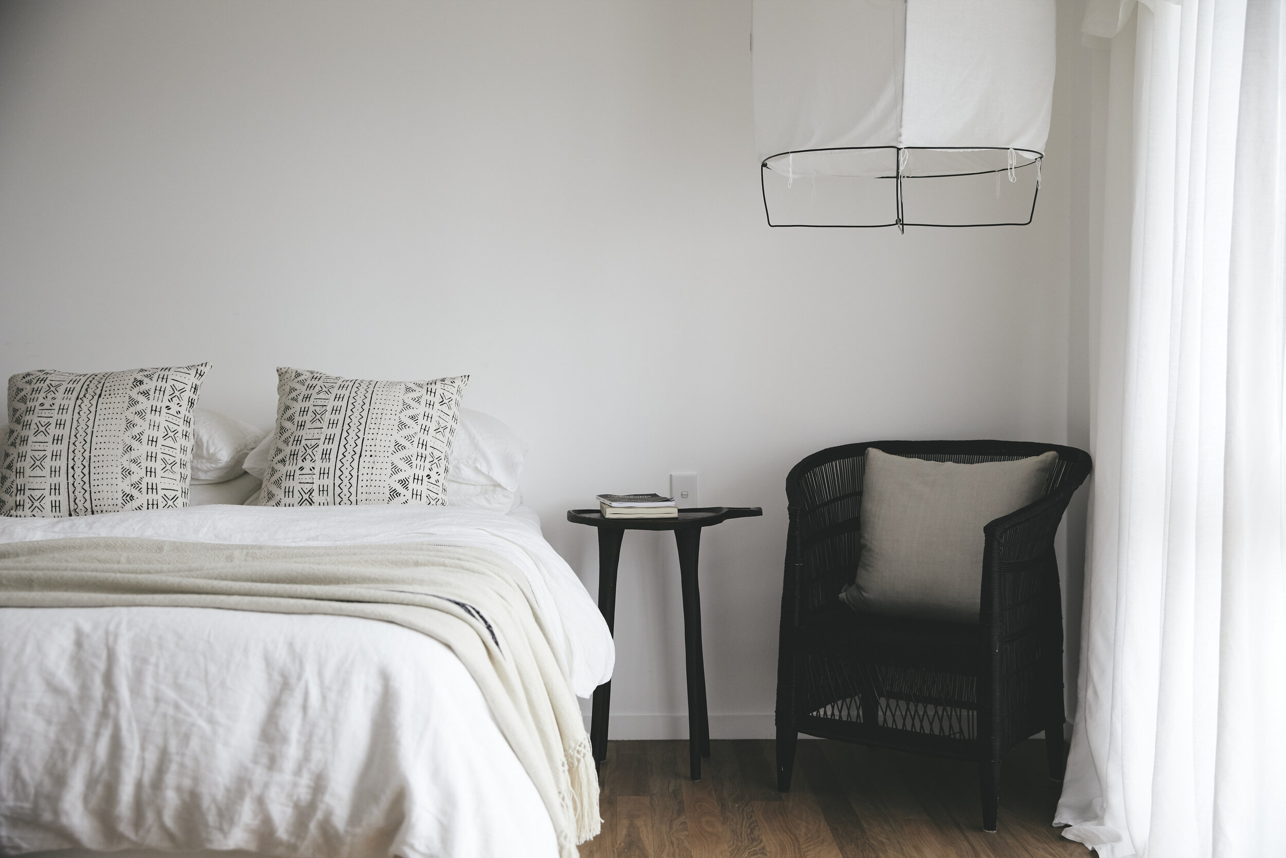 Bedroom interior with a white bed, patterned pillows, a black woven chair, and a small black table against a white wall.
