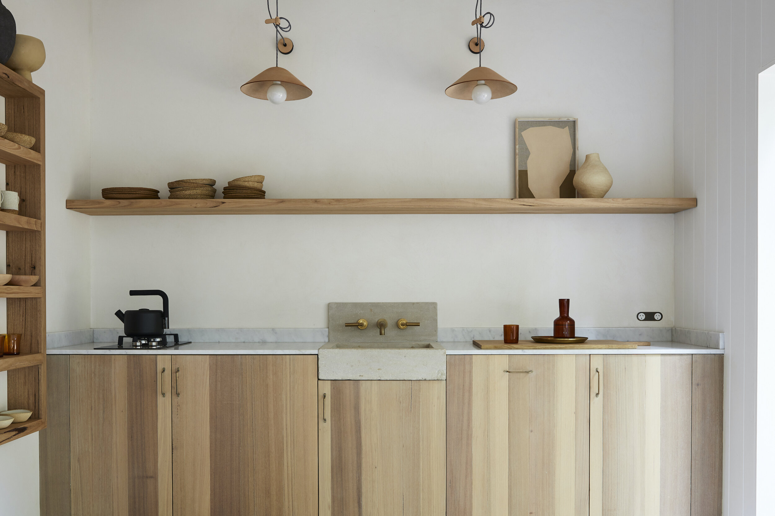 A modern kitchen interior featuring wooden cabinets, a concrete sink, brass fixtures, and minimalist decor. The space is well-lit and tidy.