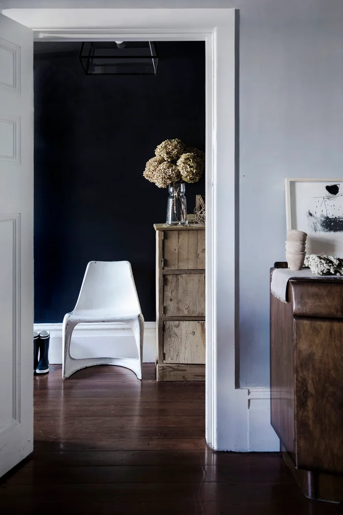Interior view through a white doorway showing a white chair, wooden cabinet with dried hydrangeas, and dark accent wall styled by Natalie Walton.