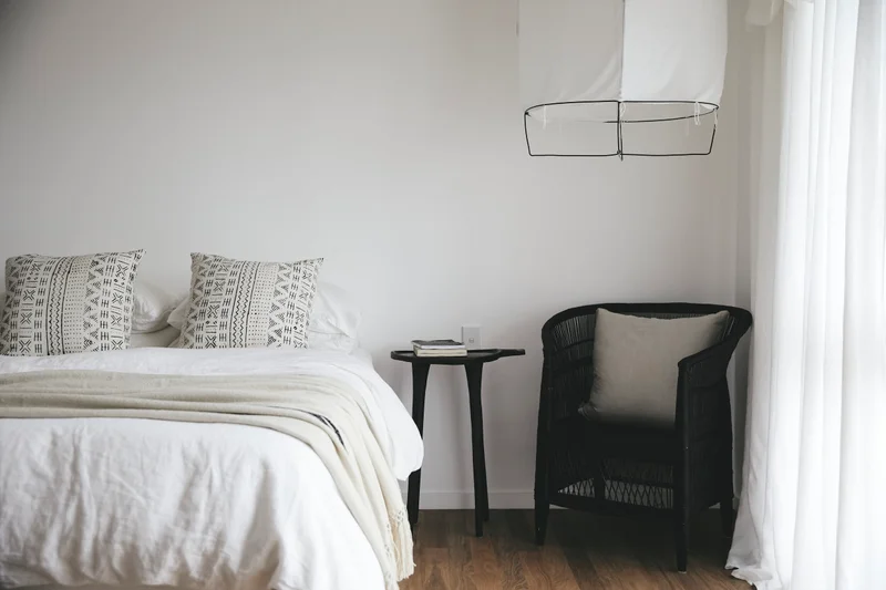 Minimalist bedroom interior styled by Natalie Walton with white bedding, black side table, and wicker armchair.