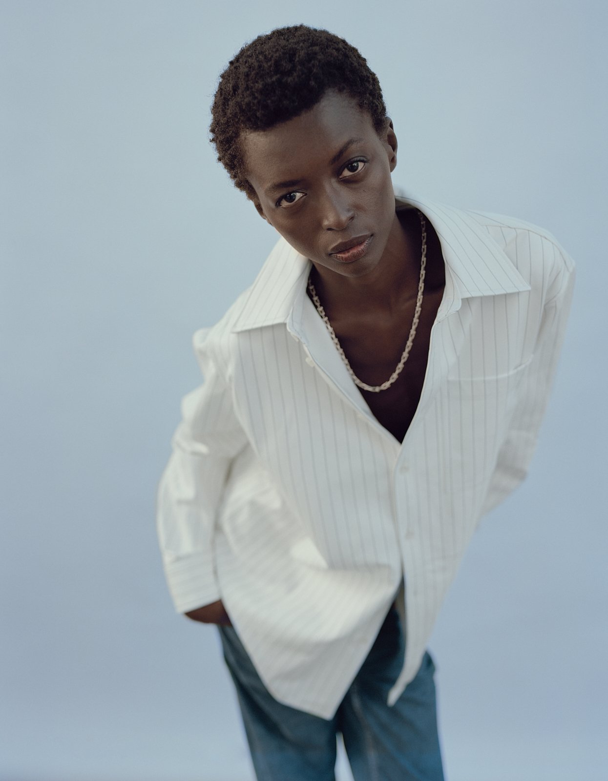 Adult Black female model in a vertical, medium shot. She wears a striped white button-down shirt and a gold necklace in a studio setting.
