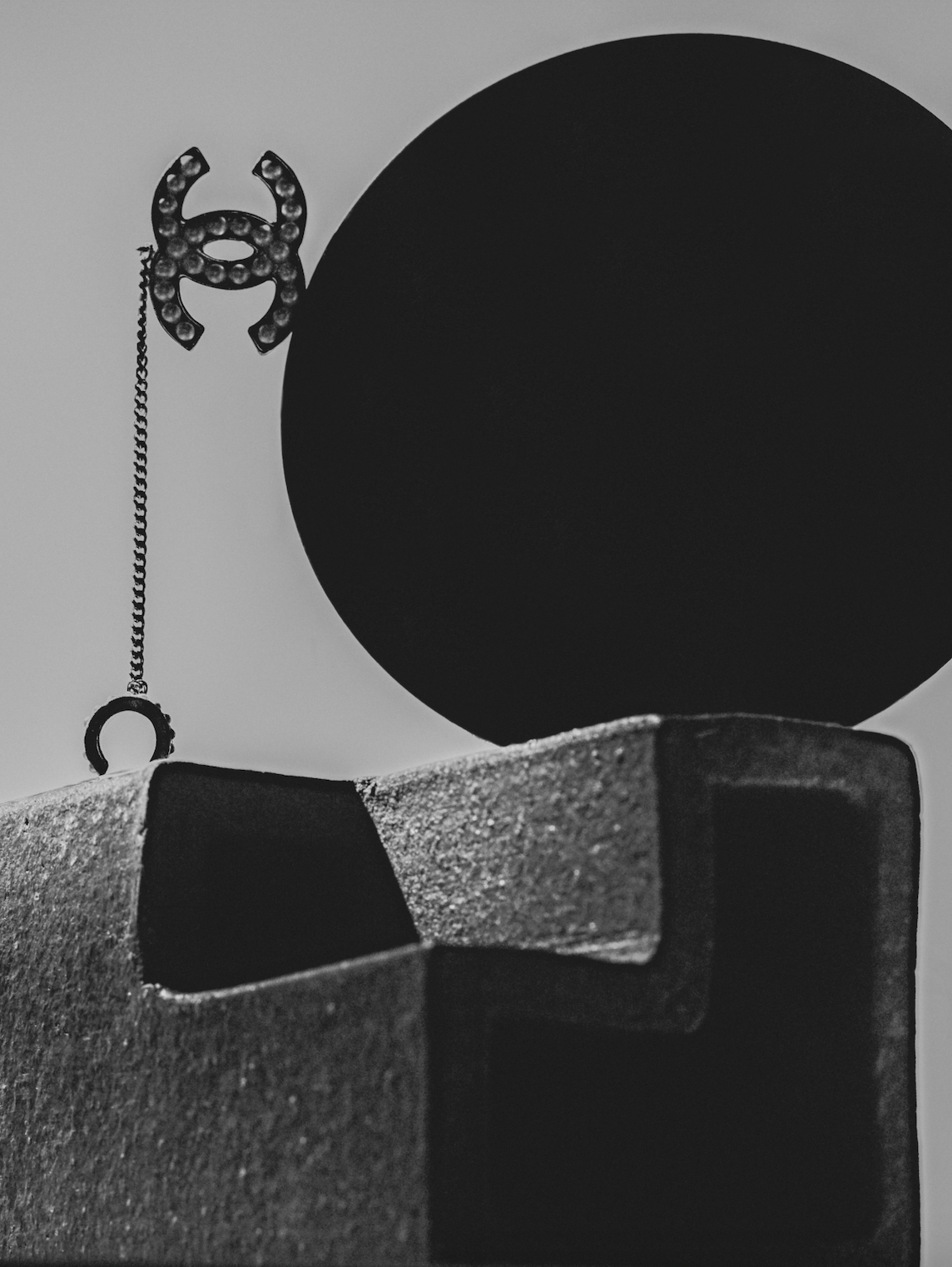 Black and white still life features a fashion brooch, black circular object and a textured block, artfully arranged.