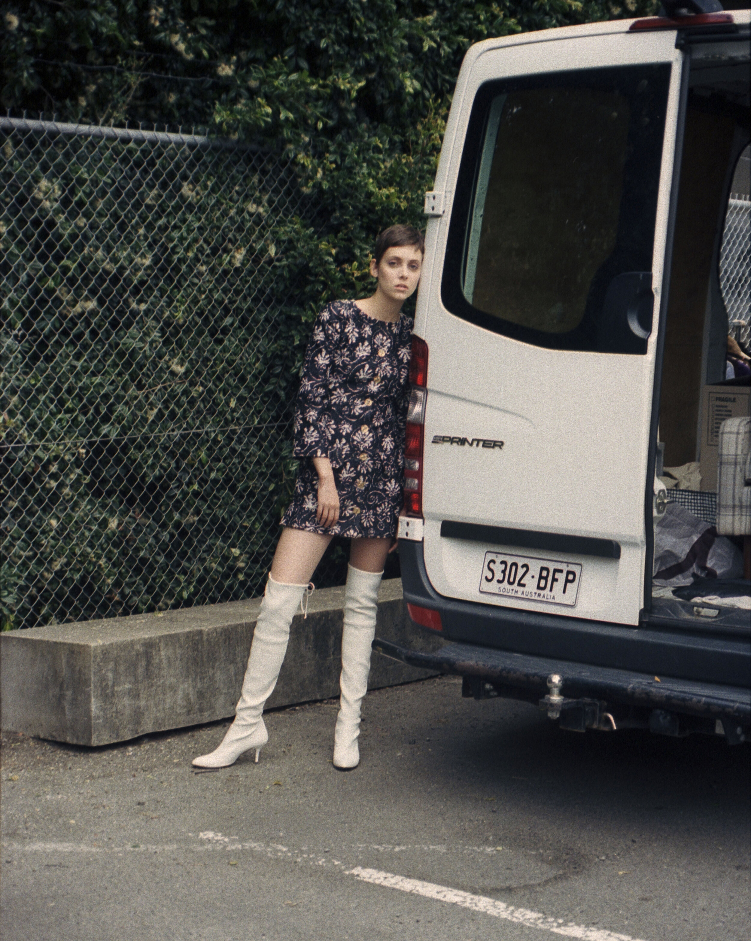 A woman in a floral dress and white boots leans against a white van, posing in an outdoor urban setting with a chain-link fence.