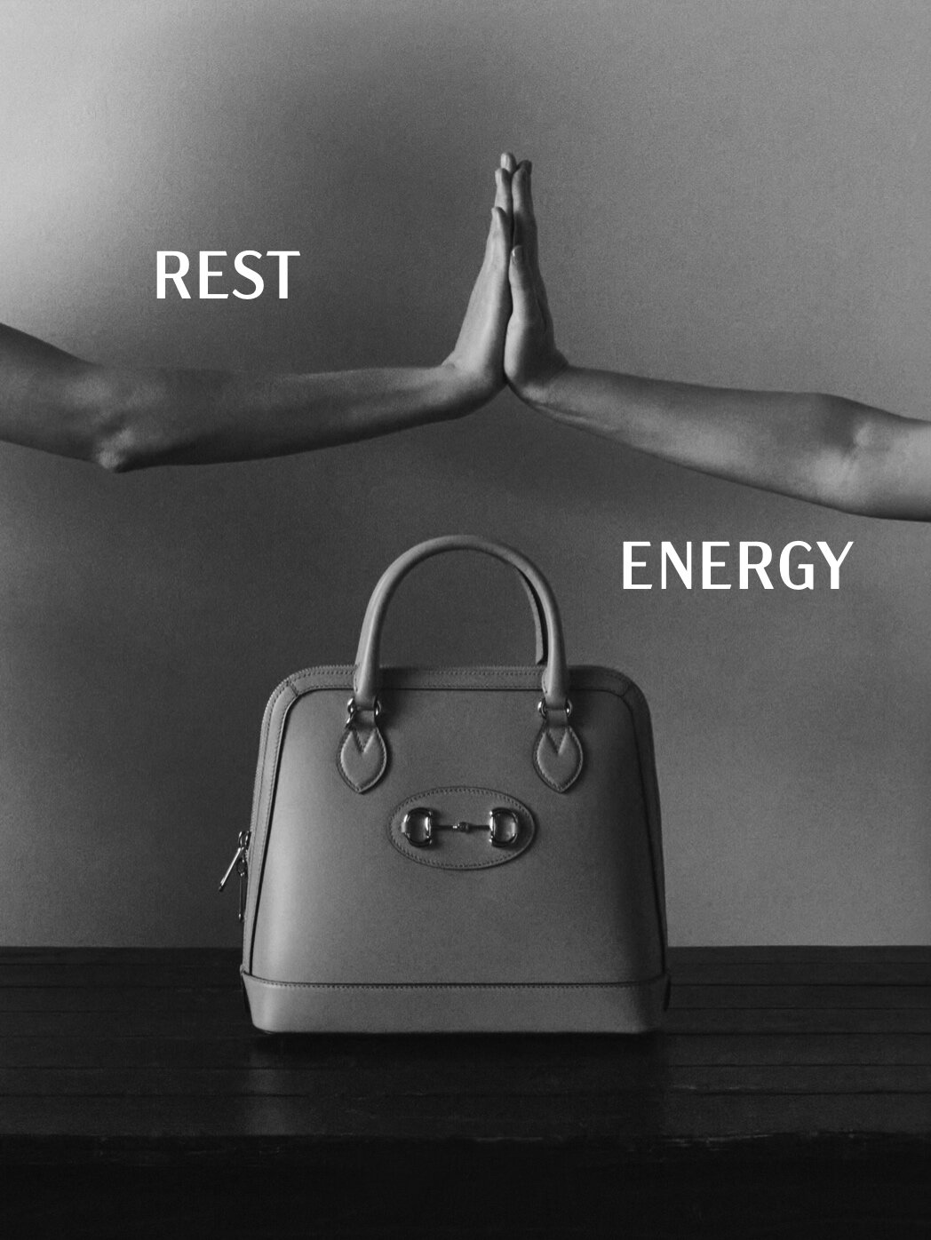 Fashion still life: dome handbag displayed on dark wood surface with two hands placed above on a gray background. Minimalist monochrome.