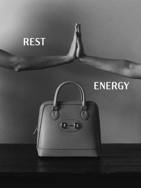 Fashion still life: dome handbag displayed on dark wood surface with two hands placed above on a gray background. Minimalist monochrome.