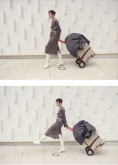 Woman in a striped dress and white boots walking with a hand truck and a box in front of a textured white wall.