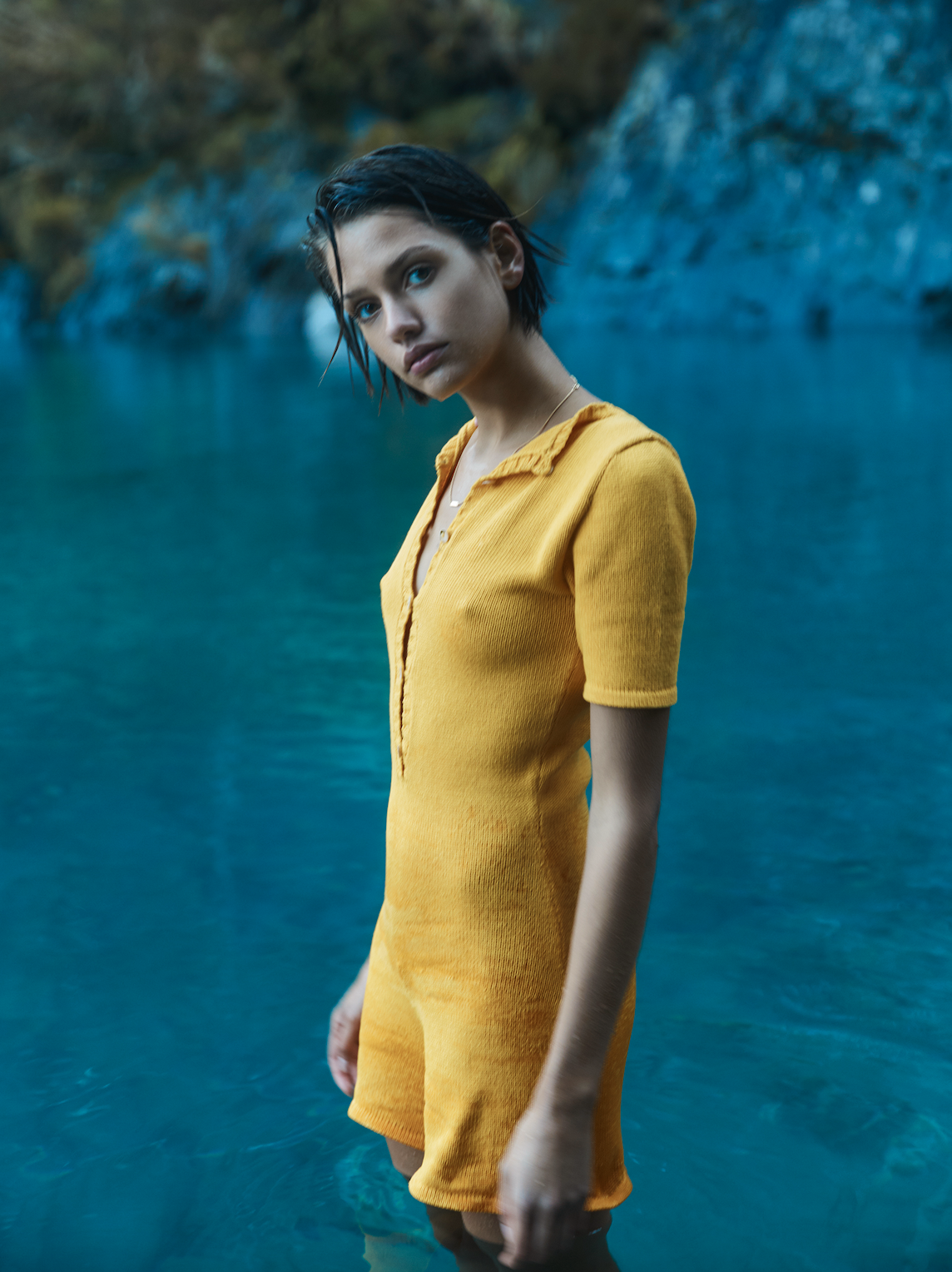 A young woman in a yellow knit romper stands waist-deep in blue water with rocks and foliage in the background, creating a serene outdoor portrait.
