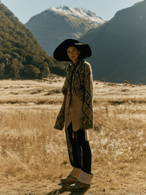 A woman in a black hat and patterned cardigan stands in a field of golden grass with mountains in the background on a sunny day.