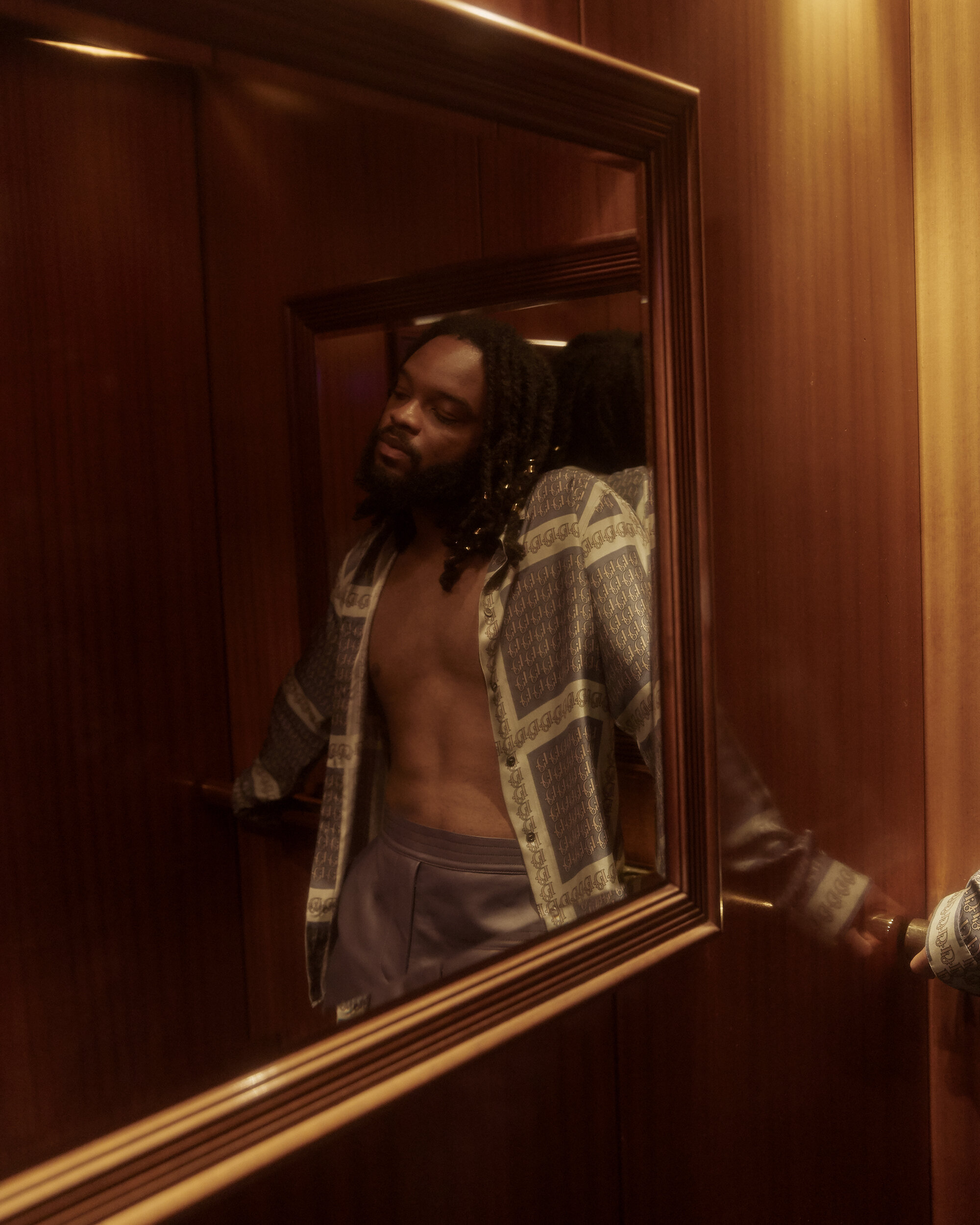 Man with dreadlocks wearing an unbuttoned patterned shirt reflected in a wood-framed mirror in a warm-toned interior