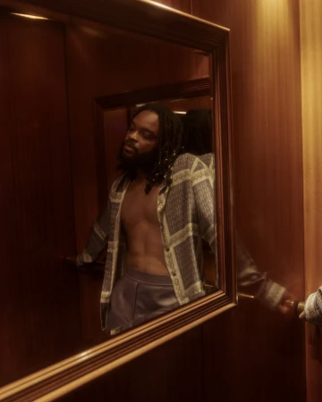 Man with dreadlocks wearing an unbuttoned patterned shirt reflected in a wood-framed mirror in a warm-toned interior