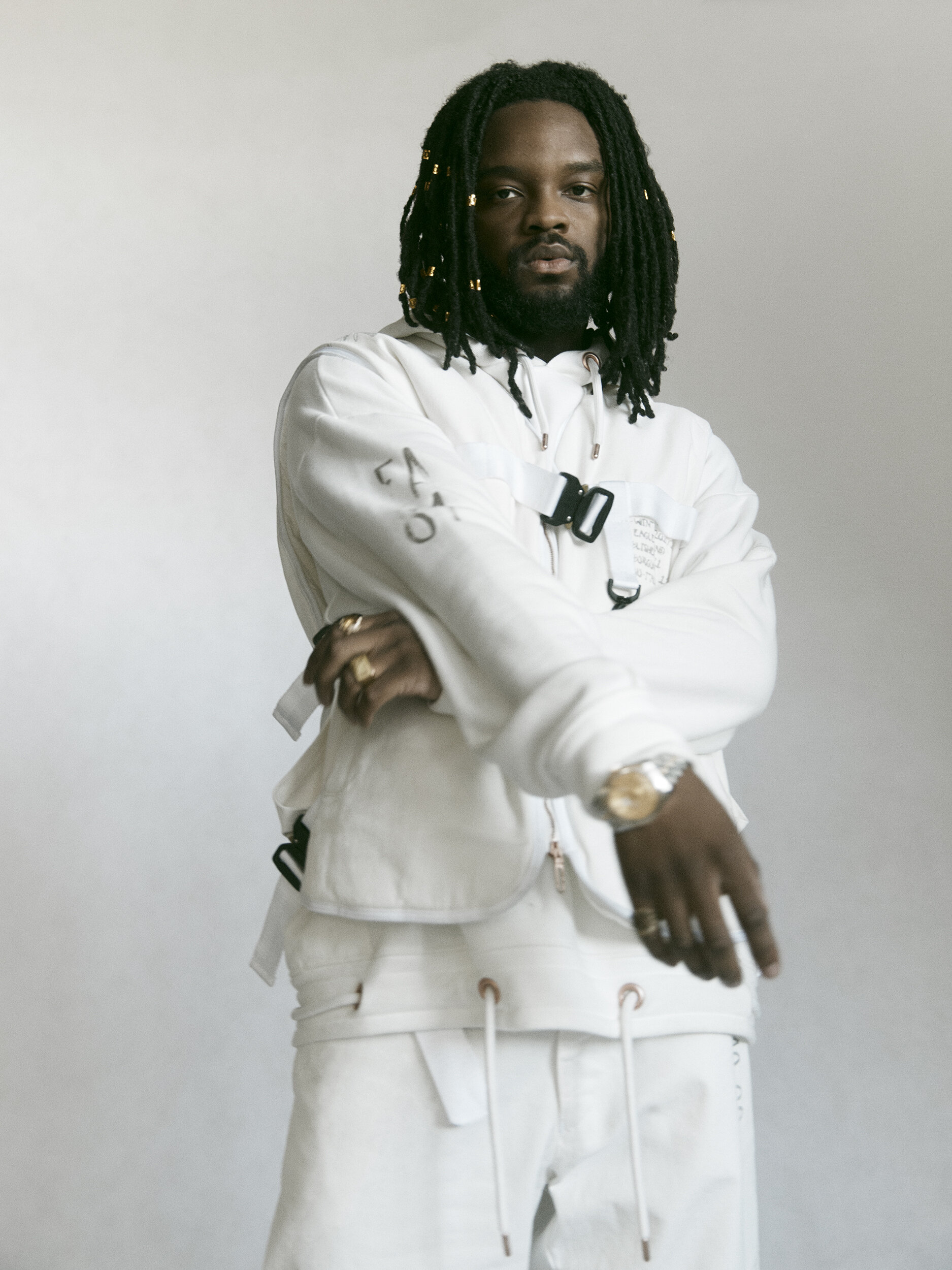 Portrait of a stylish adult male with dreadlocks wearing a white jacket and pants in a studio, posing against a white backdrop.