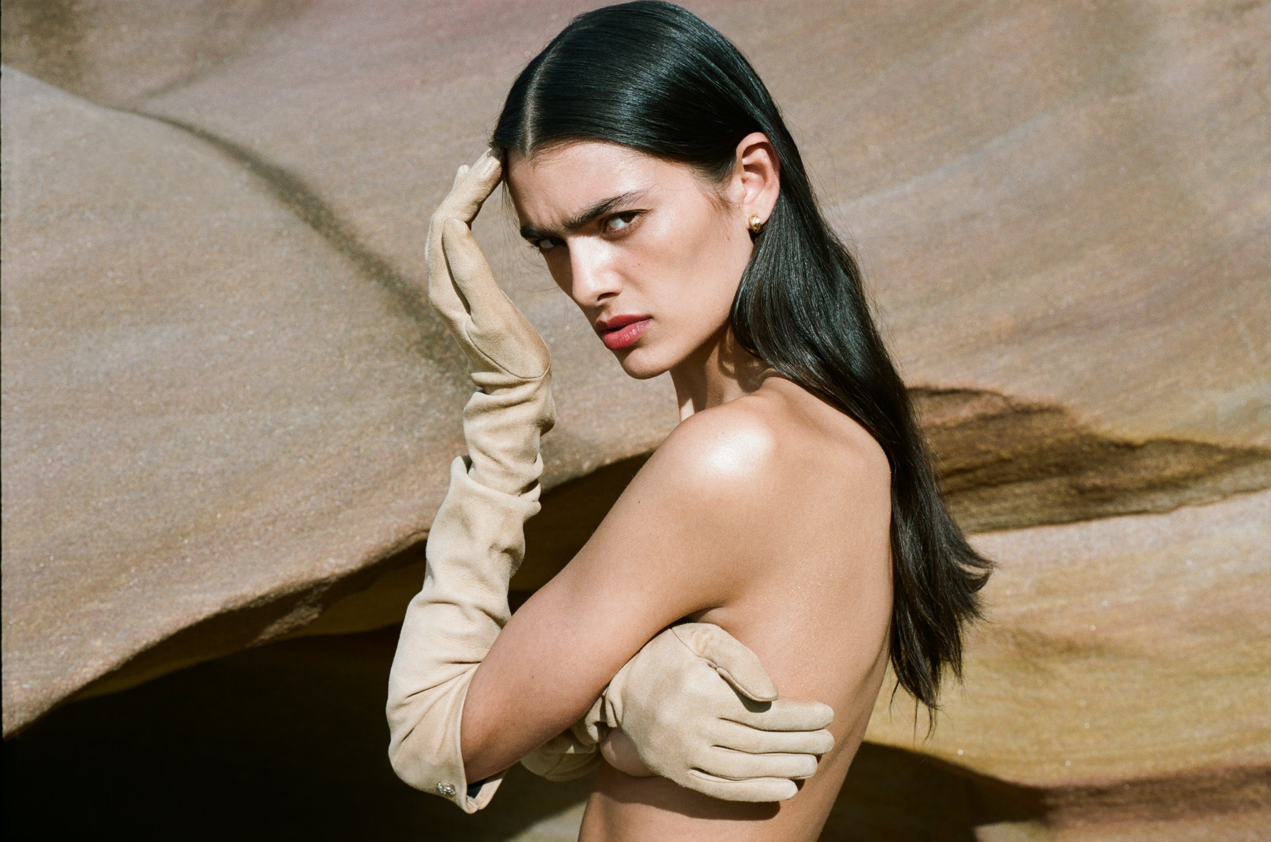 Woman with long black hair poses with light beige gloves against a neutral tan rock formation backdrop in a fashion photograph.