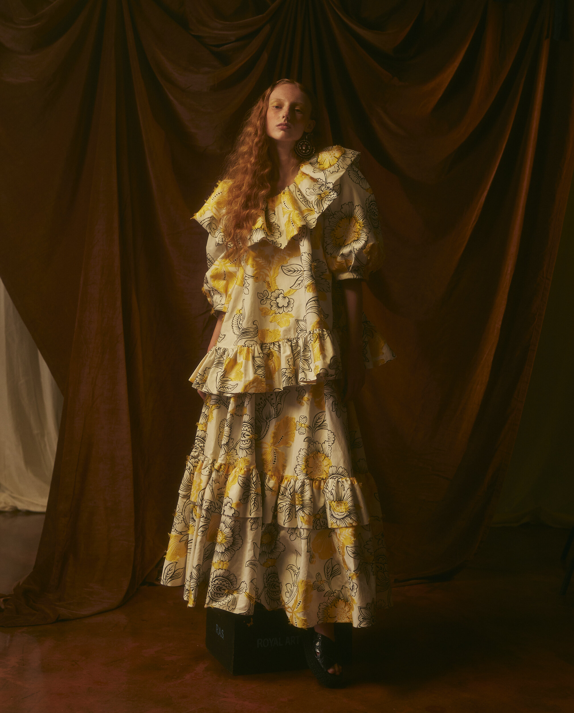 Model in a yellow and white floral print ruffled dress standing before draped brown fabric by Natalie Petrevski