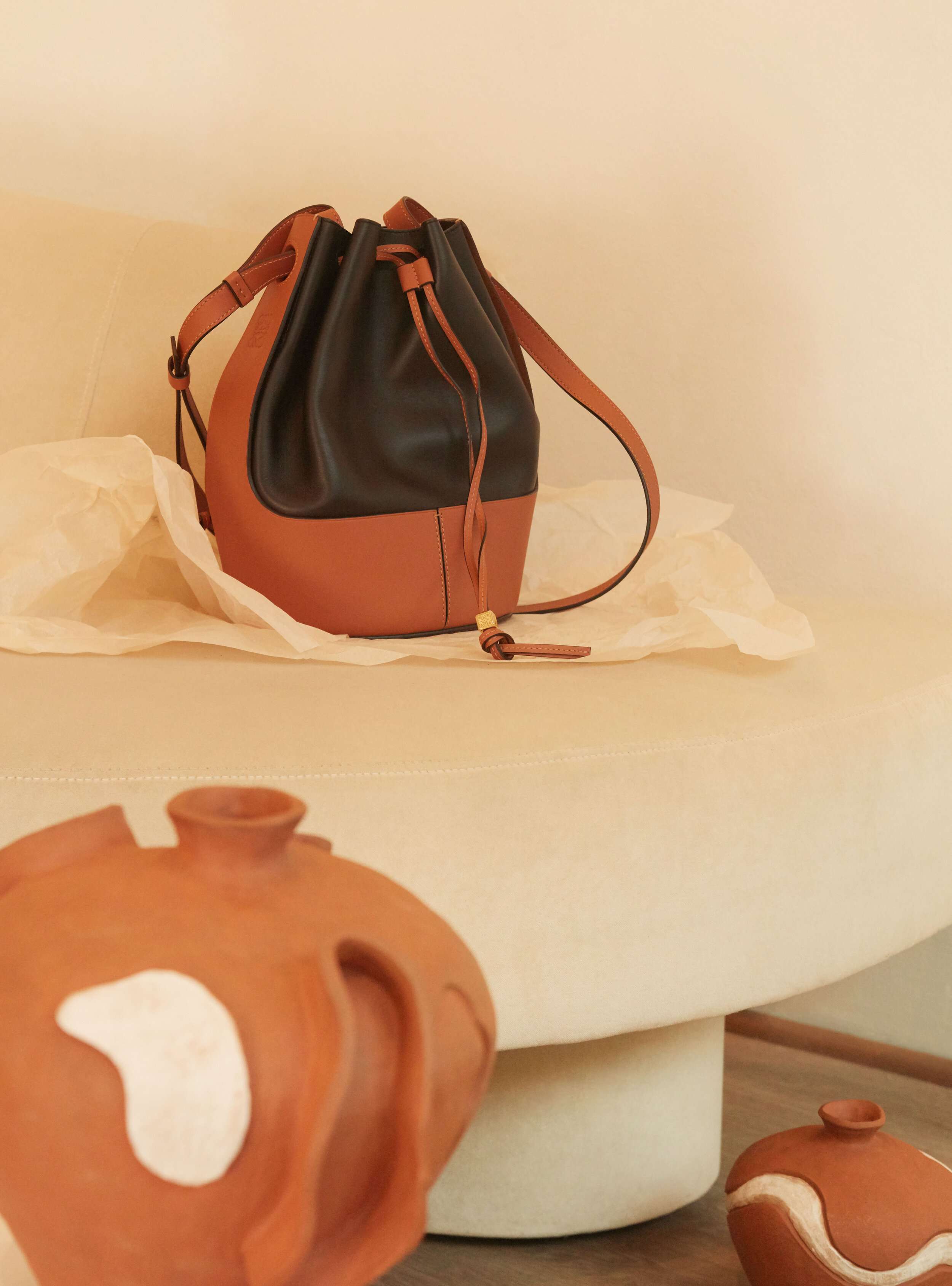 Studio shot of a brown and black leather bucket bag on a cream platform, styled with brown earthenware vases in soft, diffused light.