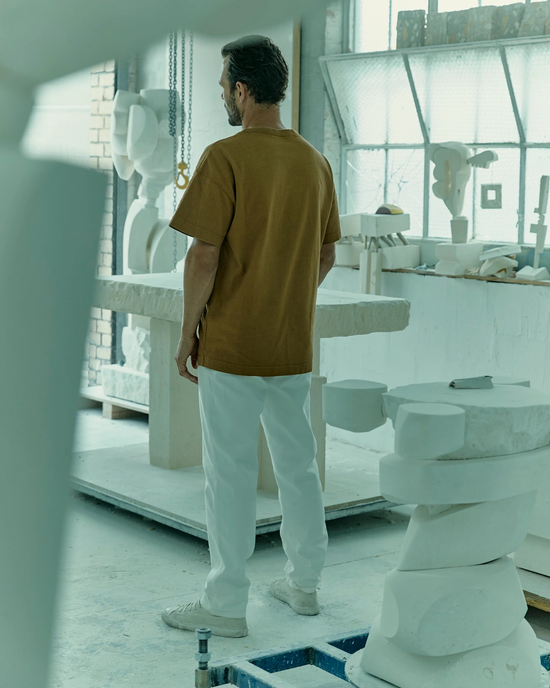 Man wearing a brown t-shirt and white trousers standing in a bright sculpture studio filled with stone artworks.