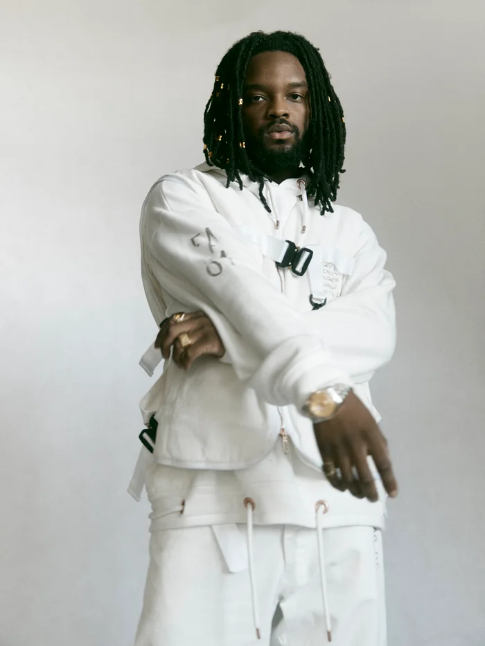 Portrait of a black man with dreadlocks wearing a white utility hoodie and pants against a seamless studio background