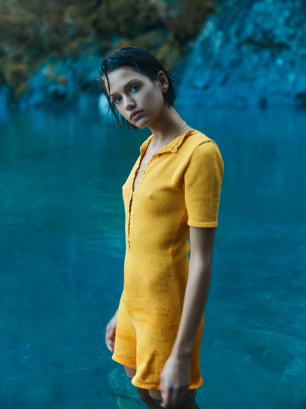 Woman with short wet hair wearing a yellow knit romper standing in a natural body of water