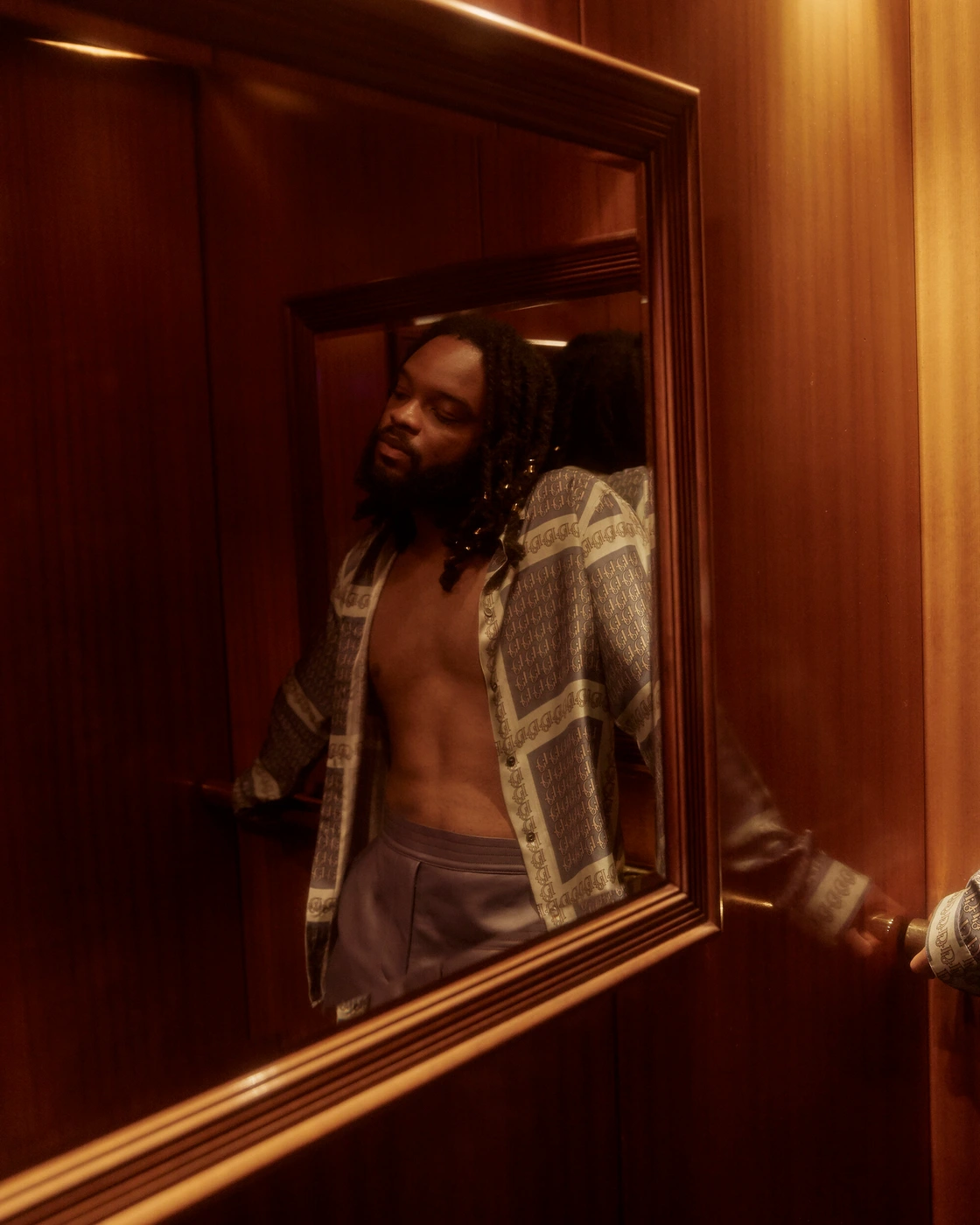 Man with dreadlocks wearing an unbuttoned patterned shirt reflected in a wood-framed mirror in a warm-toned interior