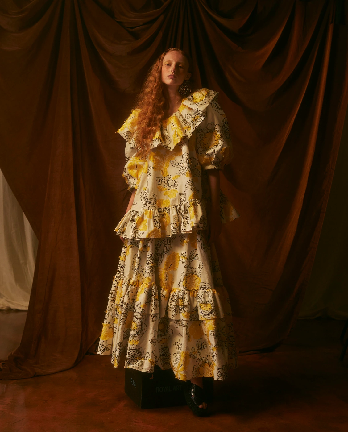 Model in a yellow and white floral print ruffled dress standing before draped brown fabric by Natalie Petrevski