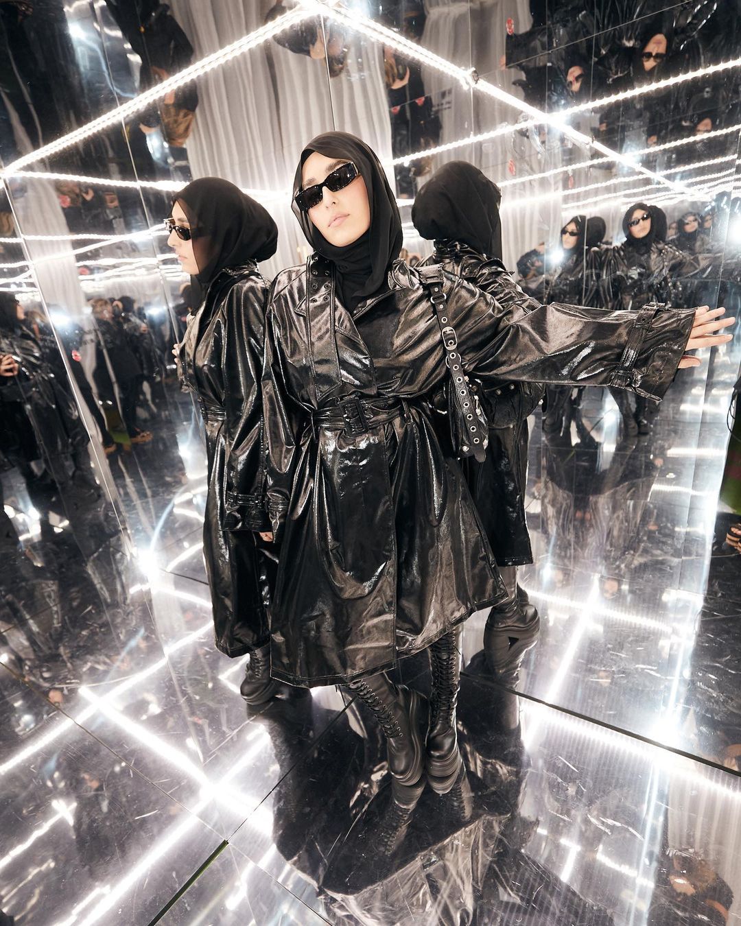 Woman in black hijab and shiny trench coat stands in mirrored infinity room with neon lights. Fashion-forward modest style.