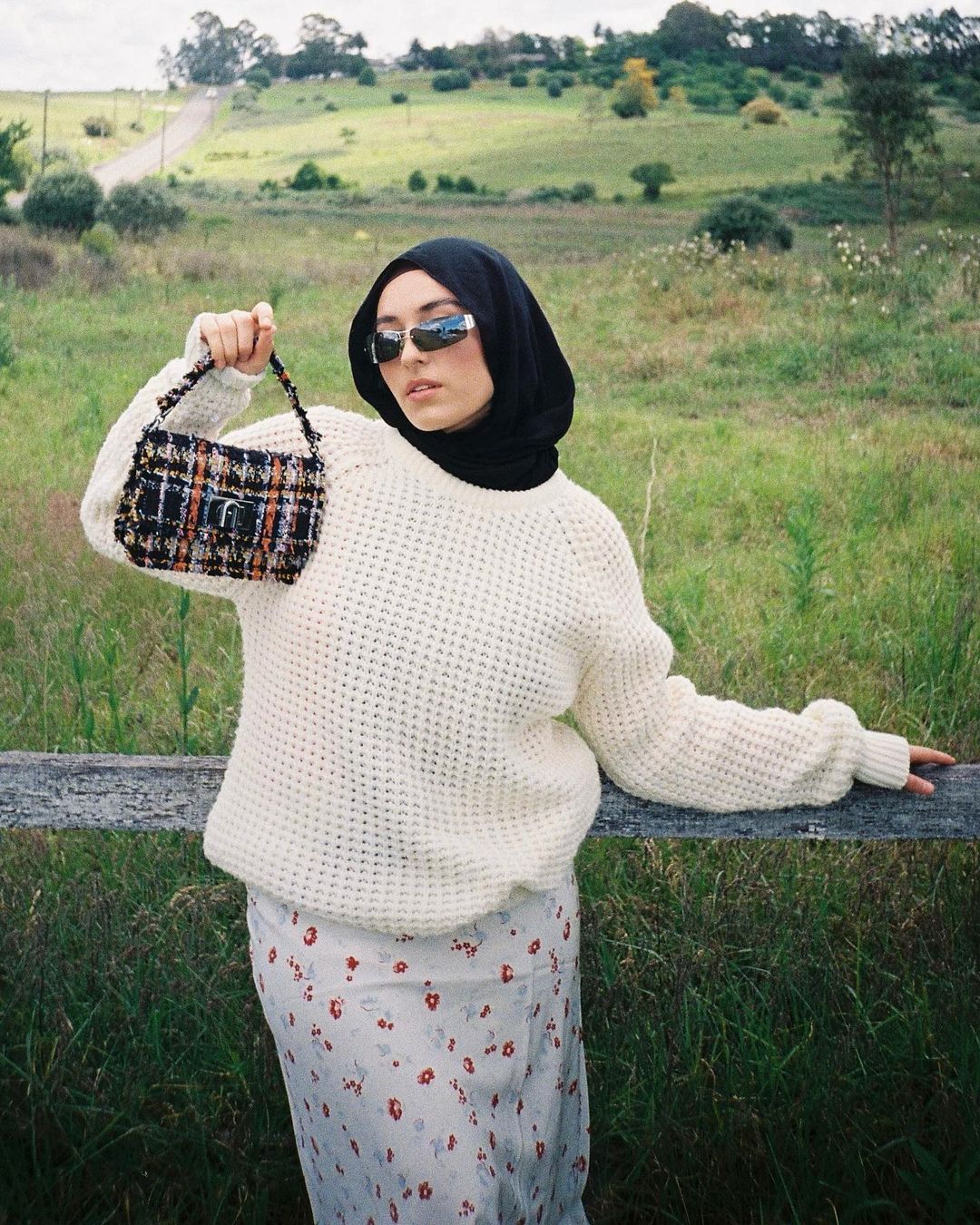 Adult woman wearing a hijab and sunglasses posing in a field with a designer handbag and floral dress near a wooden fence.