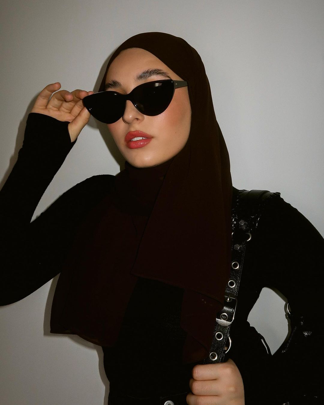 A woman wearing a brown hijab and black sunglasses poses in front of a neutral wall with a black top and black purse.