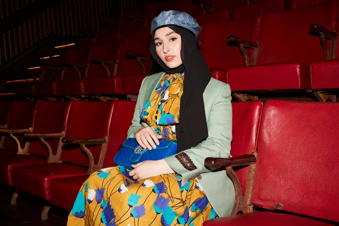 Woman wearing a hijab, green blazer, floral dress, and blue beret seated in a red theatre seat