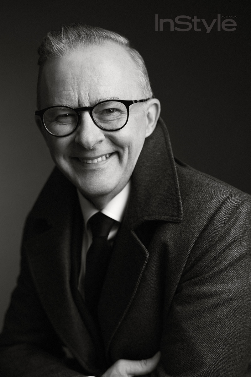Black and white portrait of a smiling mature adult man with glasses, wearing a coat and tie, against a dark background.