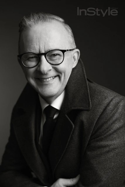 Black and white portrait of a smiling mature adult man with glasses, wearing a coat and tie, against a dark background.