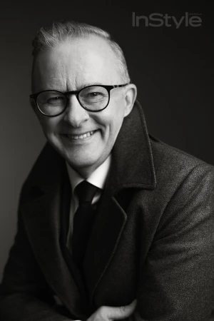 Black and white portrait of a smiling mature adult man with glasses, wearing a coat and tie, against a dark background.