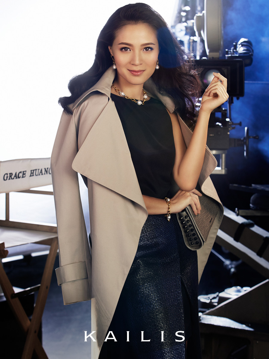 Elegant woman poses in a beige trench coat and dark skirt in front of a film set with a vintage camera and movie equipment.
