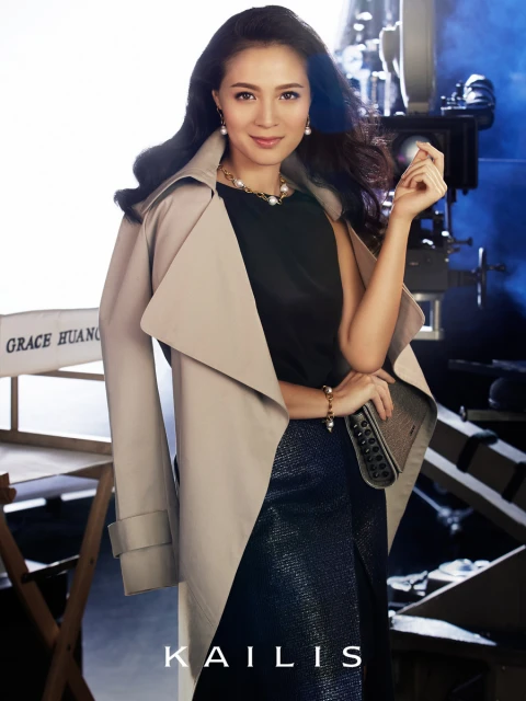 Elegant woman poses in a beige trench coat and dark skirt in front of a film set with a vintage camera and movie equipment.