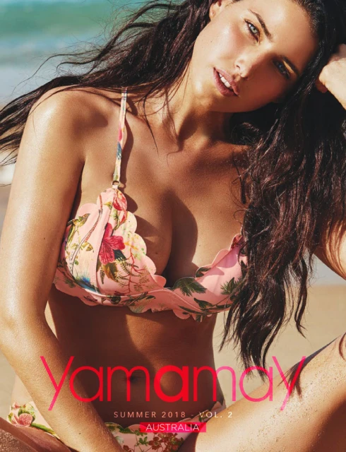 Brunette woman poses in floral bikini top on a beach, with ocean and sky visible in the background. Fashion, beauty, and summer vibe.