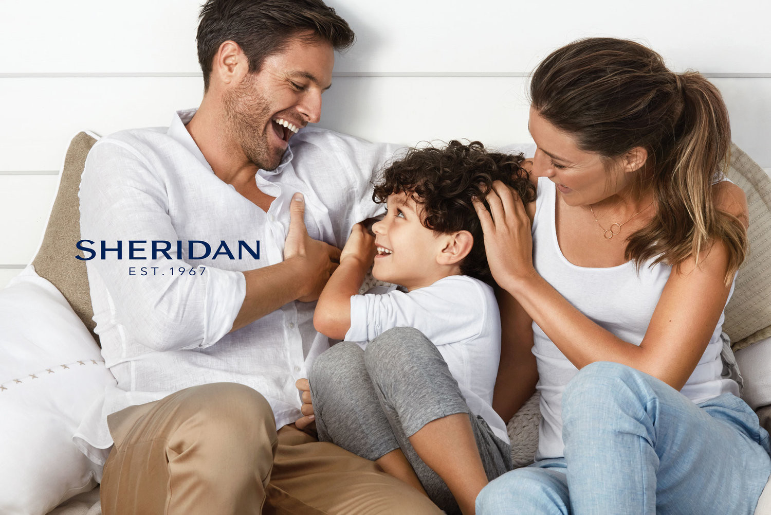 Smiling family of three relaxes together indoors; father, mother, and child sit closely while casually dressed in a bright, airy scene.