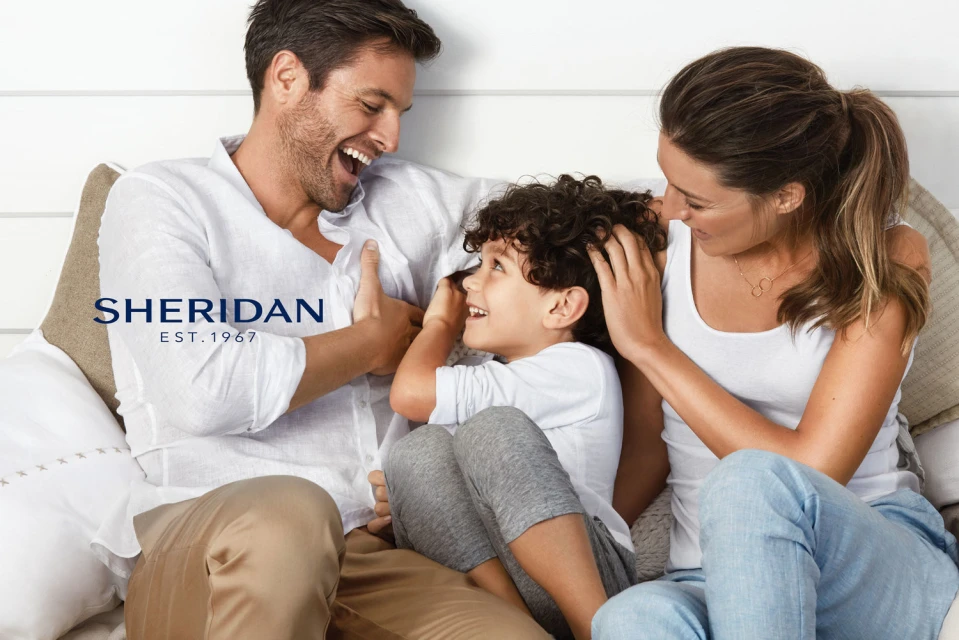 Smiling family of three relaxes together indoors; father, mother, and child sit closely while casually dressed in a bright, airy scene.