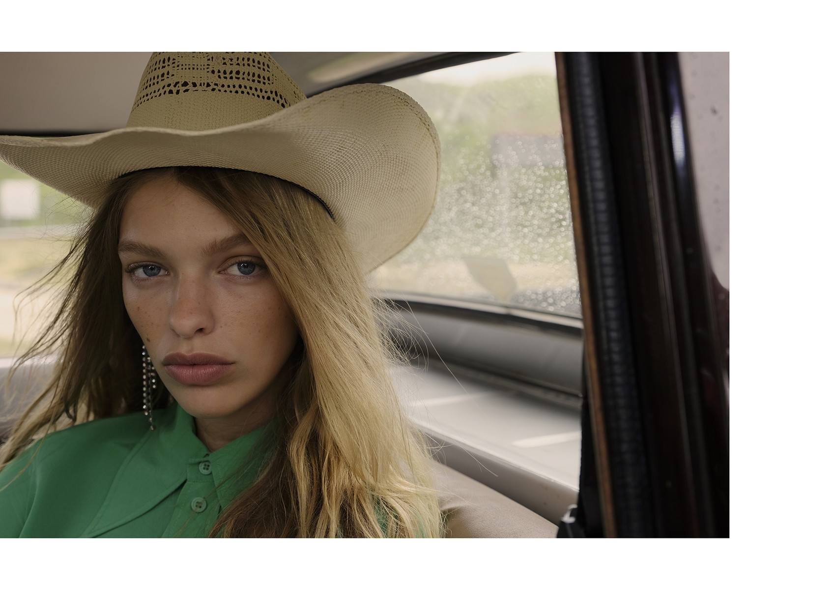 A woman with blonde hair and a cowboy hat sits in a car, looking forward with a neutral expression, with a rainy window behind her.