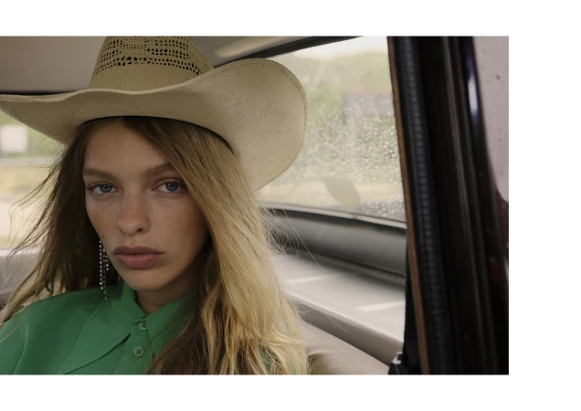 A woman with blonde hair and a cowboy hat sits in a car, looking forward with a neutral expression, with a rainy window behind her.