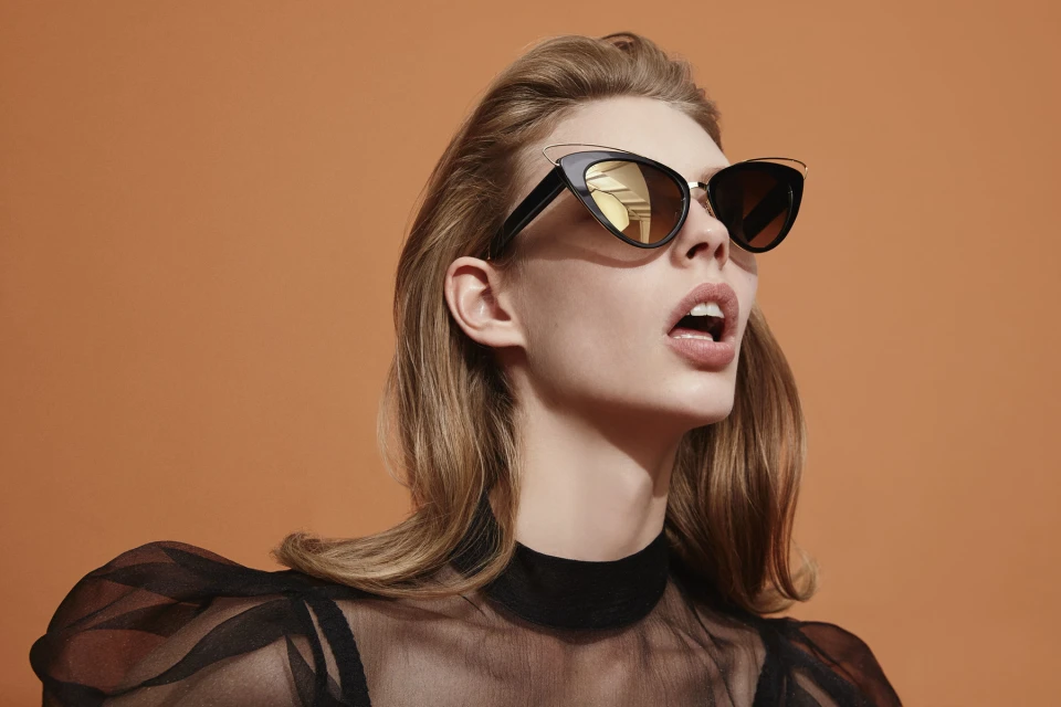 Glamorous woman with blond hair and cat-eye sunglasses poses against an orange background in sheer black top for fashion editorial.