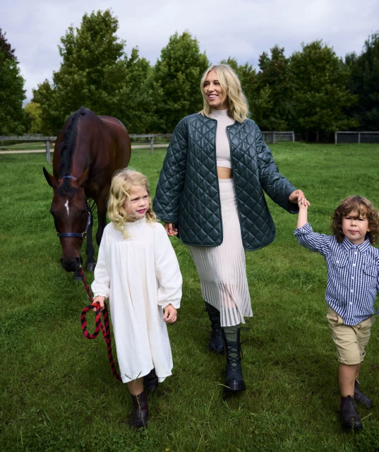 Woman with two kids walking a horse in a grassy field. Adult female holds hands with children as she walks a horse with a lead.