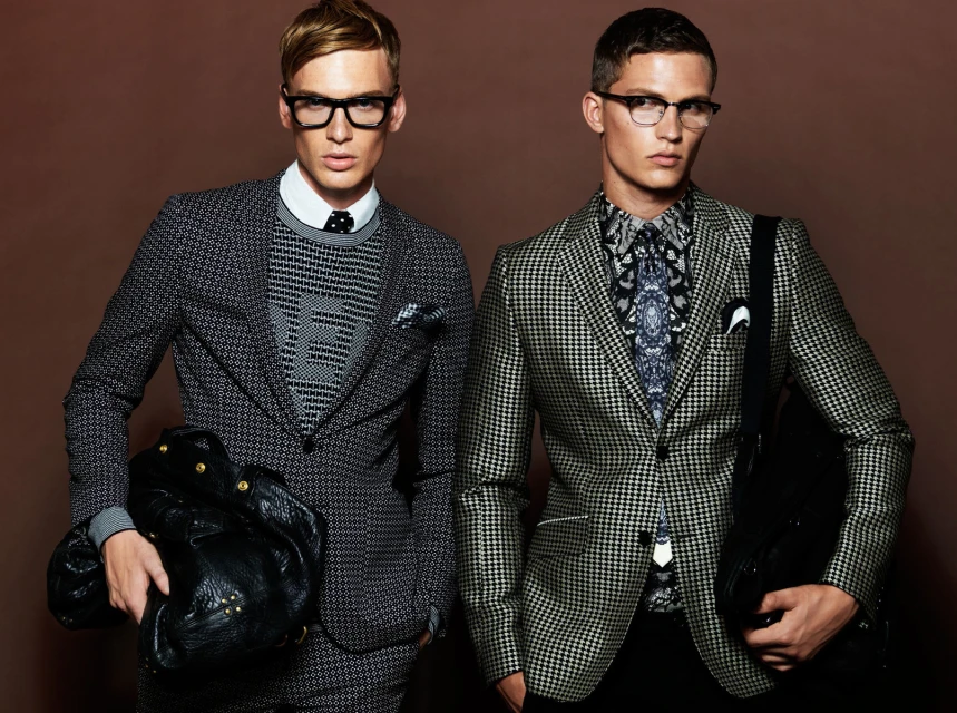 Two male fashion models pose in patterned suits and eyeglasses against a brown backdrop, showcasing menswear and stylish formalwear.