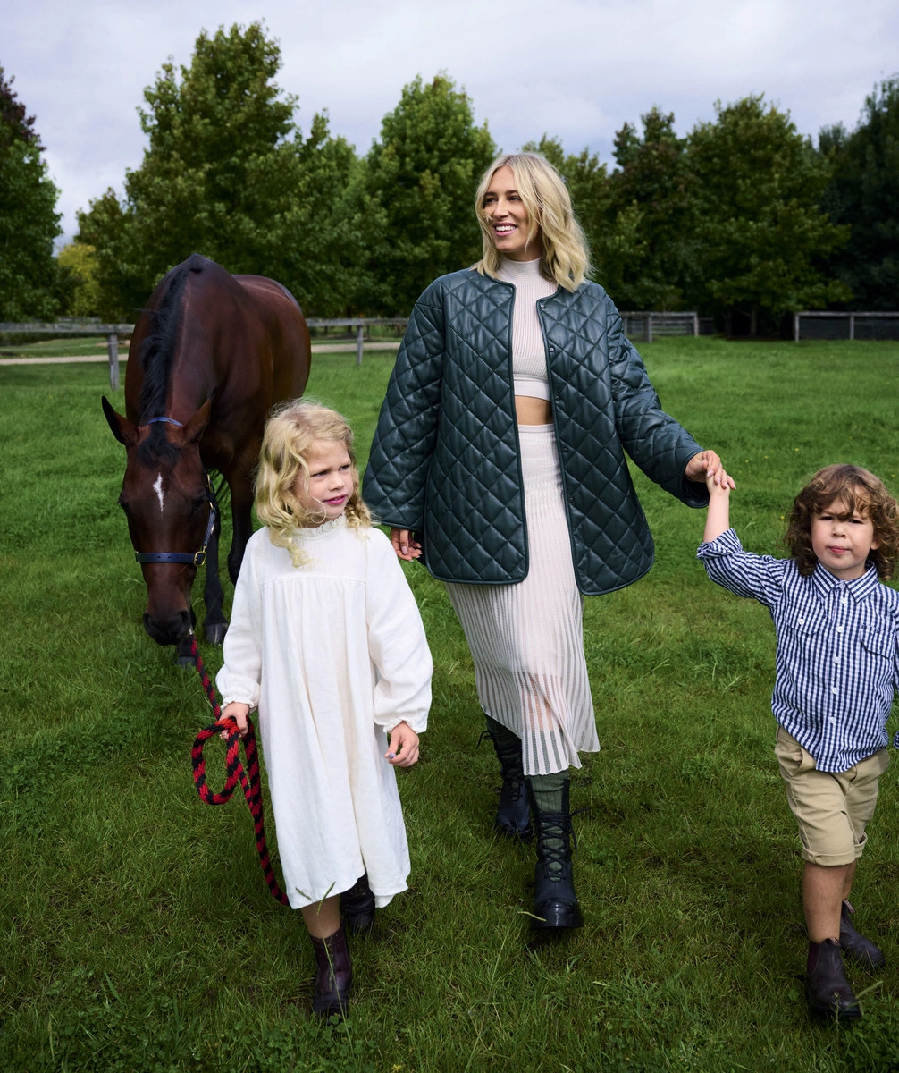 Woman in dark green quilted jacket and pleated skirt walking in a grassy field with two children and a horse