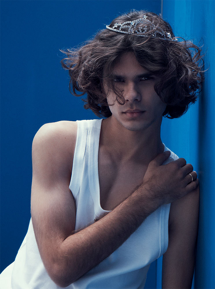 Portrait of a young adult man with long hair wearing a tiara and white tank top against a blue background.