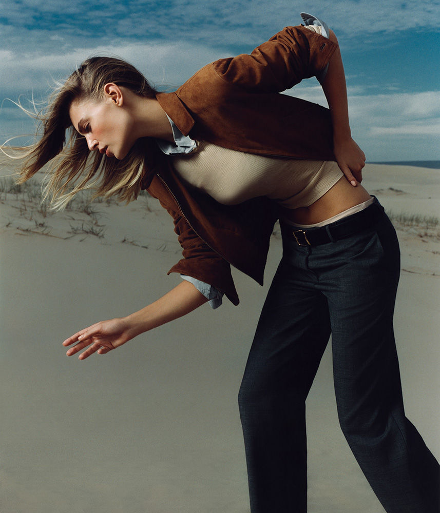 Fashion photography of a woman in a brown jacket, beige top, and black pants posing on sand dunes against a blue and cloudy sky.