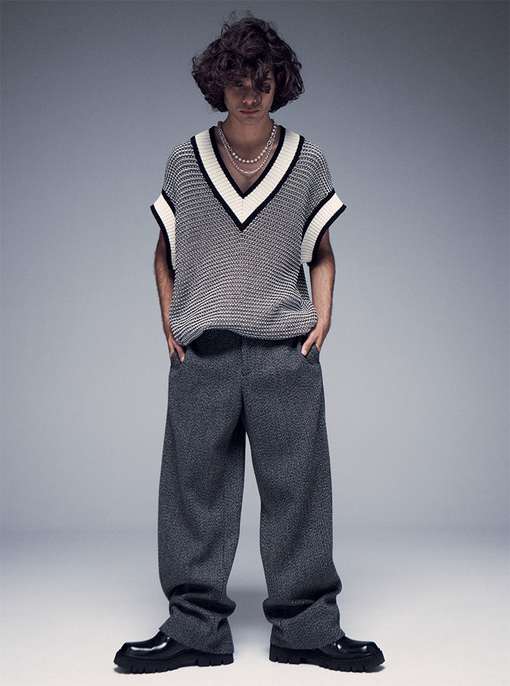 A young adult male with curly hair poses in a V-neck sweater vest, gray tweed trousers, and black lug-sole loafers against a gray backdrop.