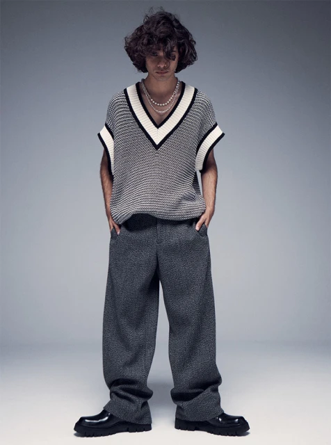 A young adult male with curly hair poses in a V-neck sweater vest, gray tweed trousers, and black lug-sole loafers against a gray backdrop.