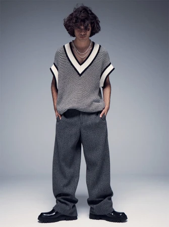 A young adult male with curly hair poses in a V-neck sweater vest, gray tweed trousers, and black lug-sole loafers against a gray backdrop.