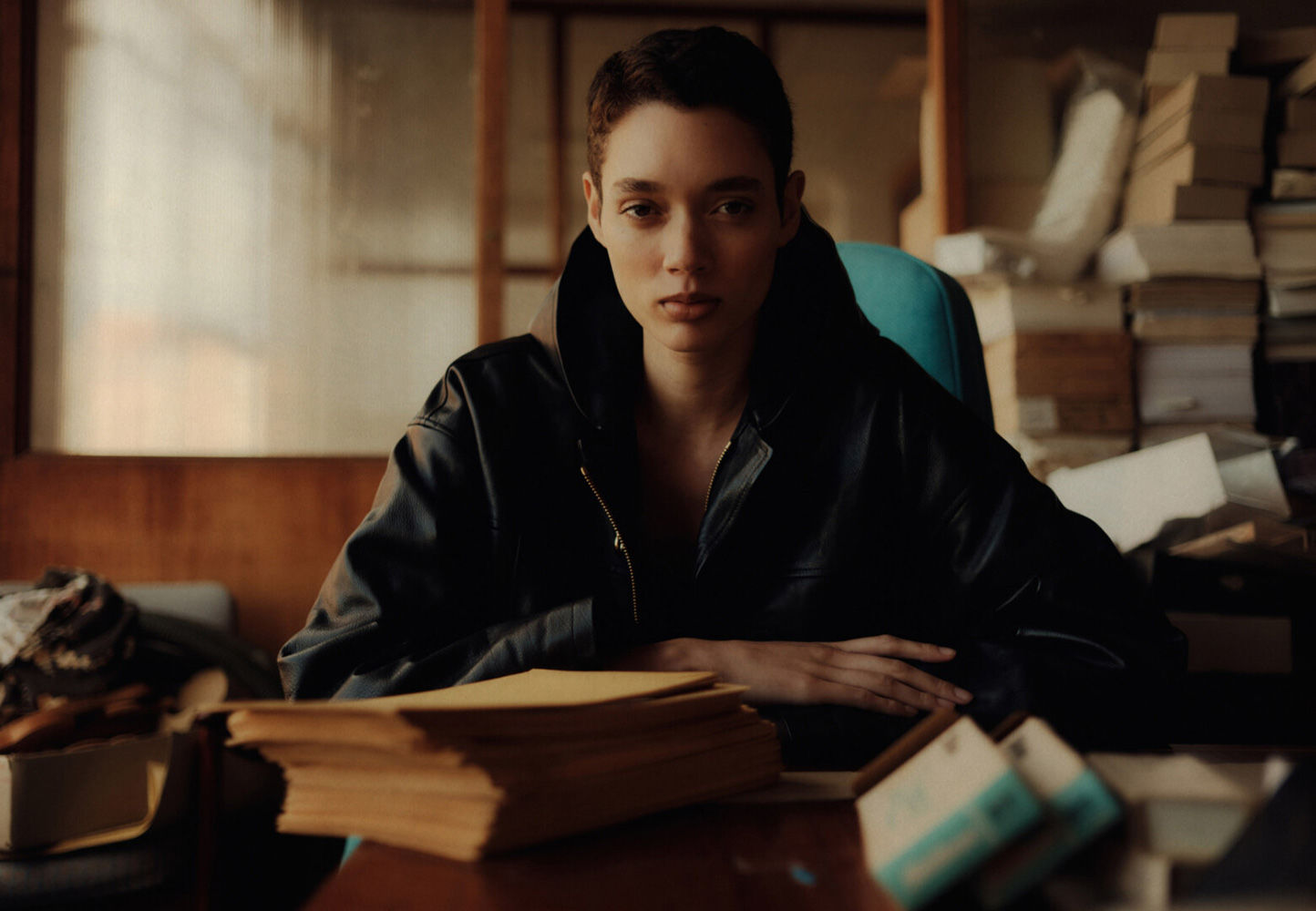 Brunette adult female in a black leather jacket sits at a wooden desk in an indoor office space with piles of books behind her.
