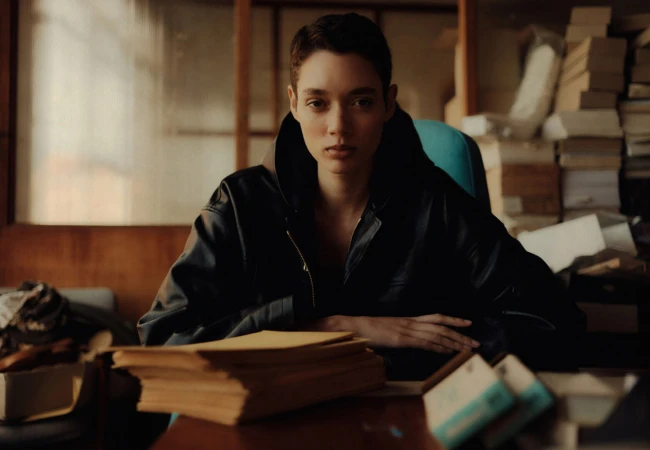 Brunette adult female in a black leather jacket sits at a wooden desk in an indoor office space with piles of books behind her.